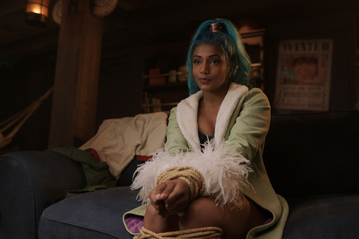 Woman with blue hair and fluffy robe sits on a cozy couch in a dimly lit room, her hands and legs tied with rope, surrounded by blankets and wooden decor; a wanted poster hangs in the background.