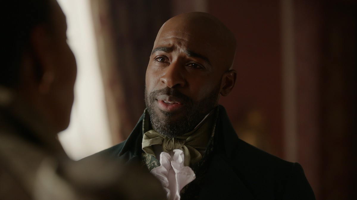 A man in historical attire with a cravat and waistcoat stands indoors in a softly lit, richly decorated room, speaking to another person whose back is to the camera.