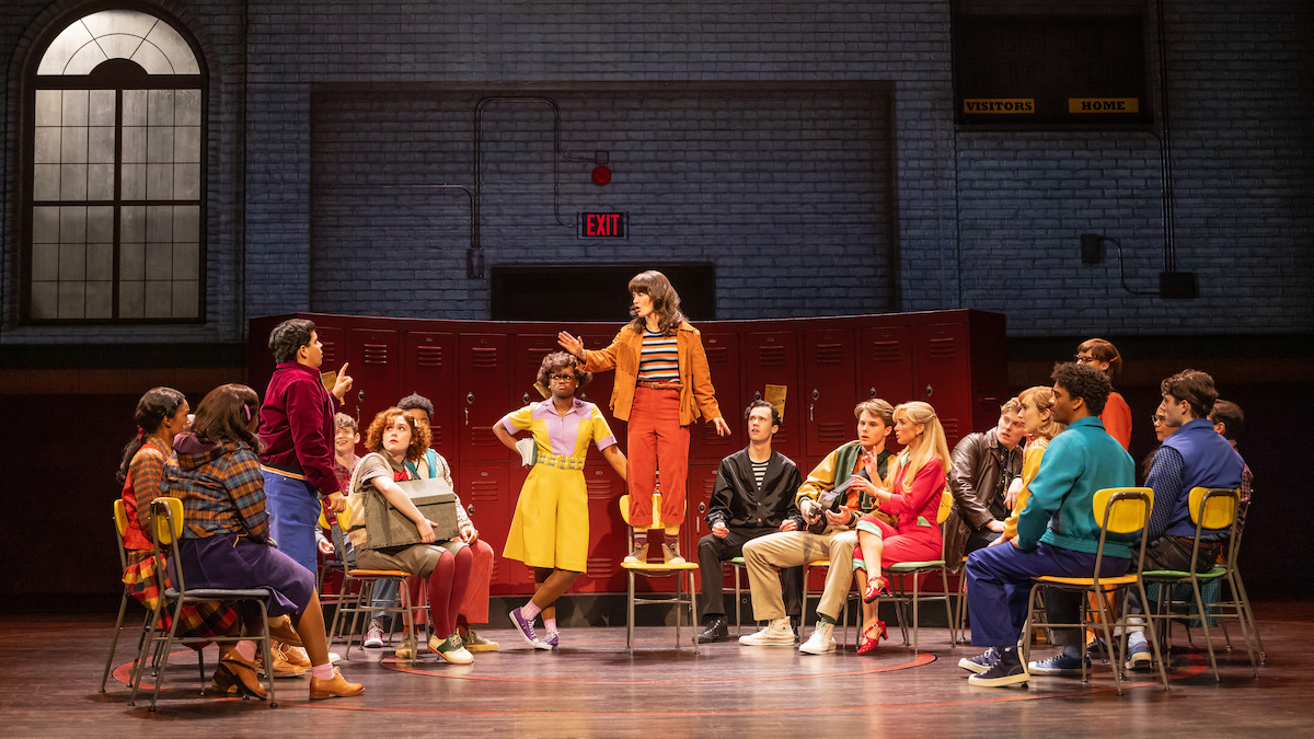 A group of people in colorful outfits sit in a circle of chairs on a stage set as a school, with one person standing and speaking passionately in the center, creating an energetic and dramatic atmosphere.