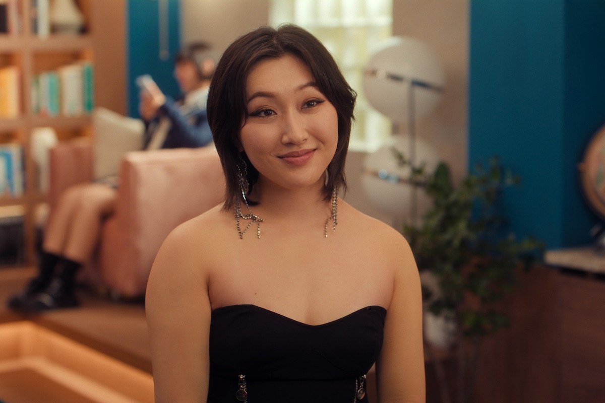 A person in a black strapless dress smiling indoors, with modern decor, bookshelves, plants, and another person blurred in the background sitting on a pink sofa.