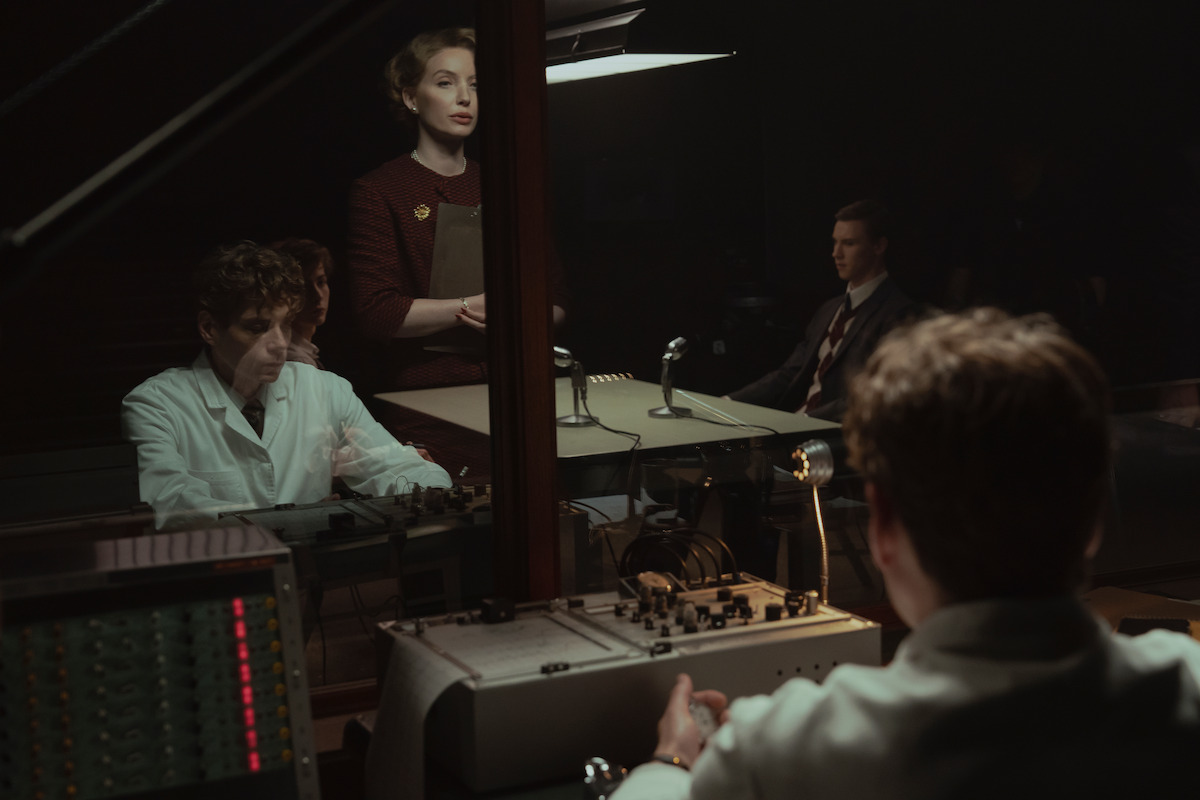A group of people in a dimly lit room, some in lab coats, appear to be conducting or observing an experiment or interrogation through glass, with vintage recording equipment and microphones on the desk.