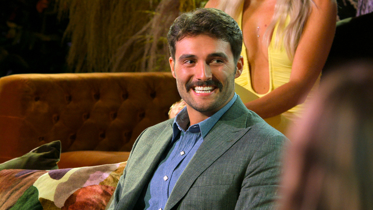 A smiling man in a gray suit sits on a brown couch in a warmly lit, cozy lounge. A woman in a yellow dress stands behind him. The atmosphere feels lively and relaxed, suggesting a social or television event.