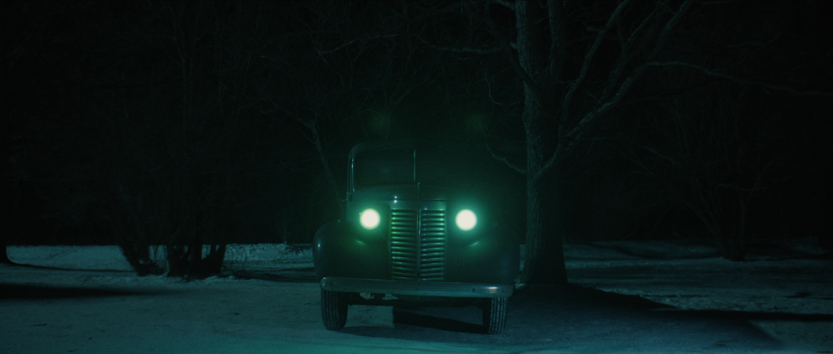 A vintage car with bright green headlights is parked on a snow-covered road at night, surrounded by leafless trees in a dark, wintry environment.