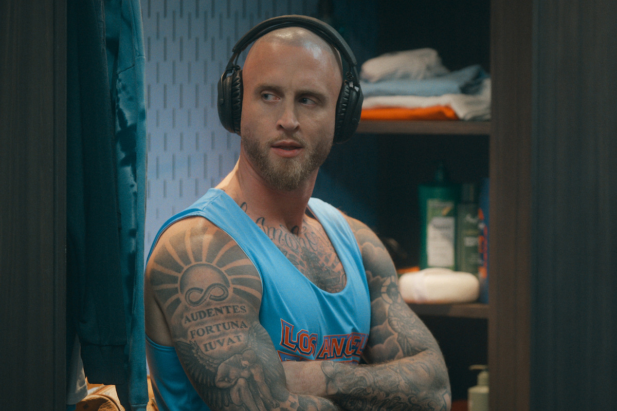 Tattooed man wearing headphones and a blue tank top stands with arms crossed in a locker room, shelves with clothes and toiletries in the background.