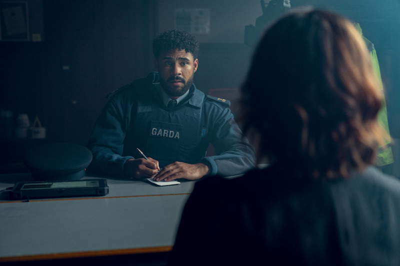 A police officer wearing a "GARDA" vest takes notes at a desk in a dimly lit office, facing a person whose back is to the camera, suggesting an interview or report situation.