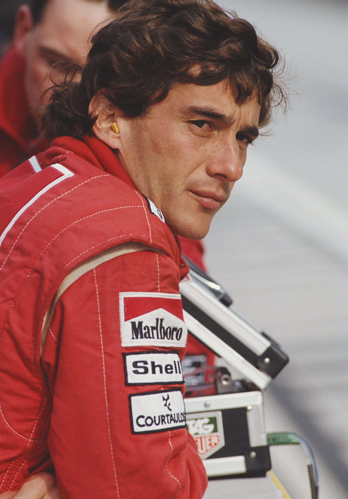 Senna wears a red racing suit and leans over a barricade at the track.