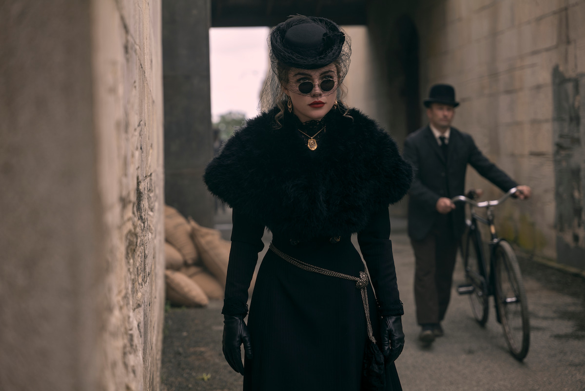 Woman in Victorian-era black attire with fur shawl and hat stands in an alleyway, wearing dark sunglasses. A man in period clothing with a bowler hat pushes a bicycle in the background beside stone walls and sandbags.