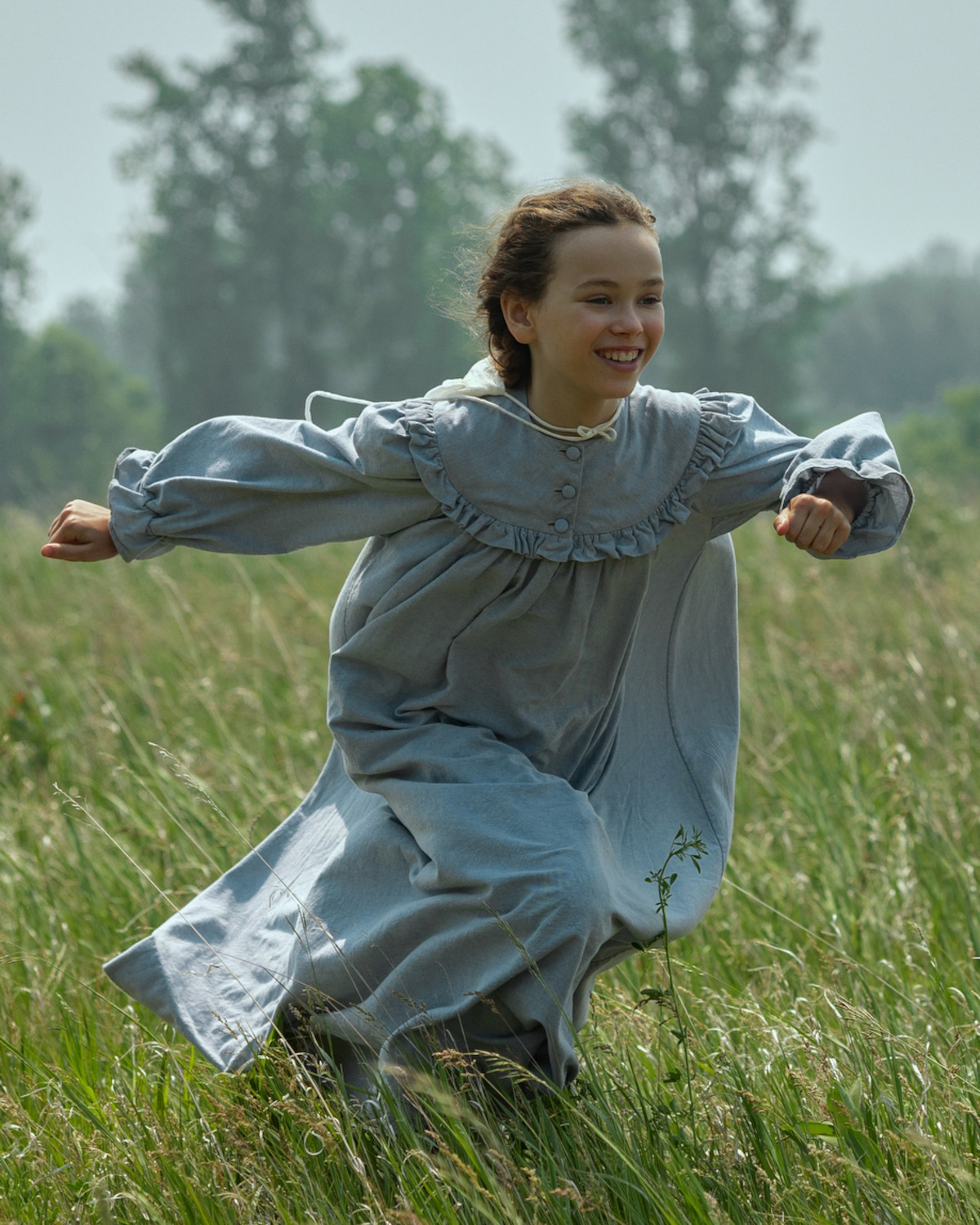 Young girl in a light blue vintage dress running joyfully through a grassy field on a sunny day, with green trees in the background and a natural, outdoor setting.