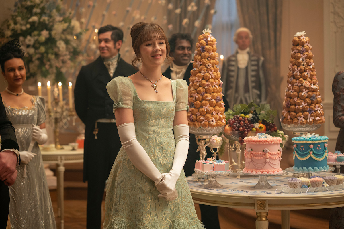 Young woman in an elegant gown stands smiling at a formal party, surrounded by guests in period attire. Ornate cakes, pastries, and elaborate desserts decorate a table in a lavish, candlelit room with floral arrangements.
