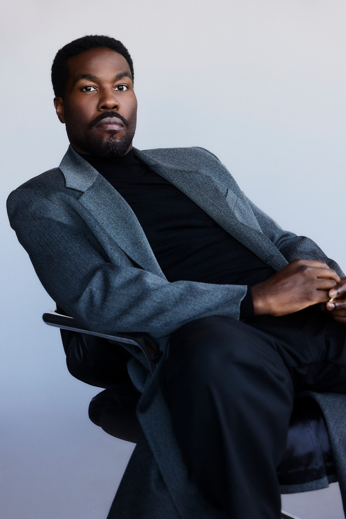 Man in a gray suit and black shirt sits in a modern chair against a plain light background, looking confidently at the camera in a professional indoor studio setting.
