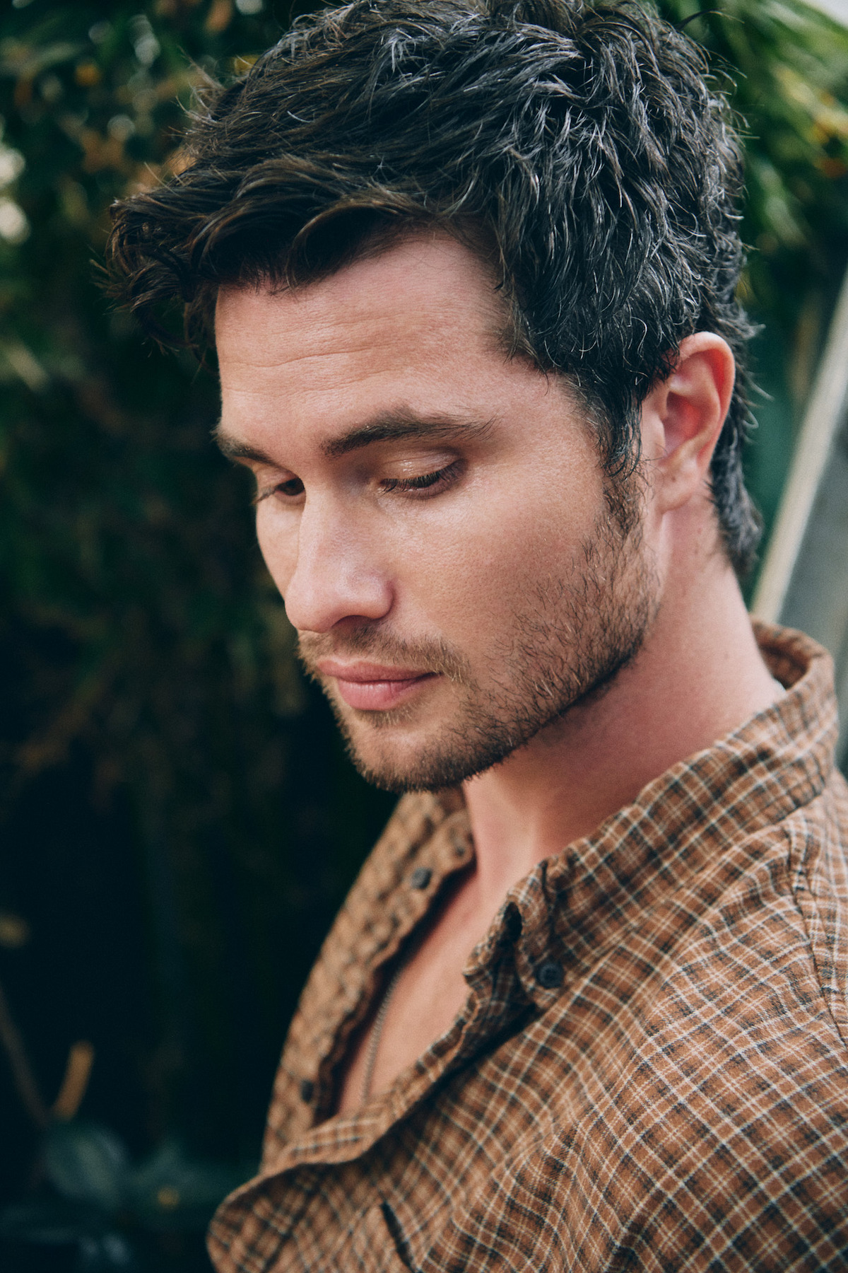 Man with dark hair and stubble wearing a brown plaid shirt looks down thoughtfully in an outdoor, lush green setting with blurred foliage in the background.
