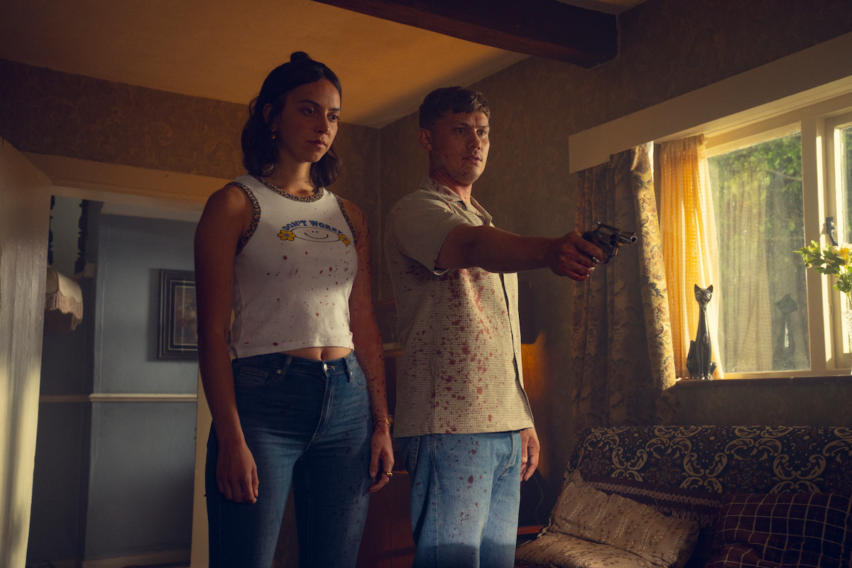 A man aiming a gun and a woman standing beside him in a dimly lit living room with sunlight streaming through the window, creating a tense and dramatic atmosphere.