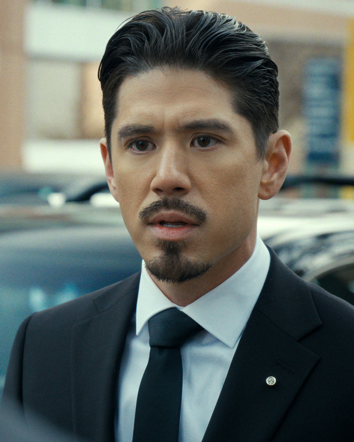 Man in a black suit and tie with slicked-back hair and mustache, standing outdoors in a parking lot with blurred cars and buildings in the background, looking serious or concerned.