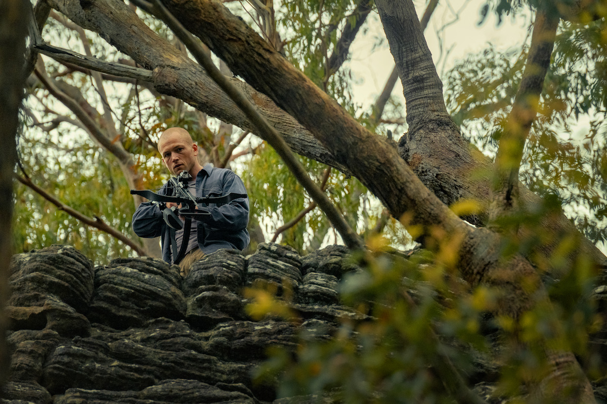 Man in outdoor military gear aiming a rifle from behind a rocky outcrop in a forested area, with tall trees and dense foliage, creating a tense, suspenseful atmosphere.