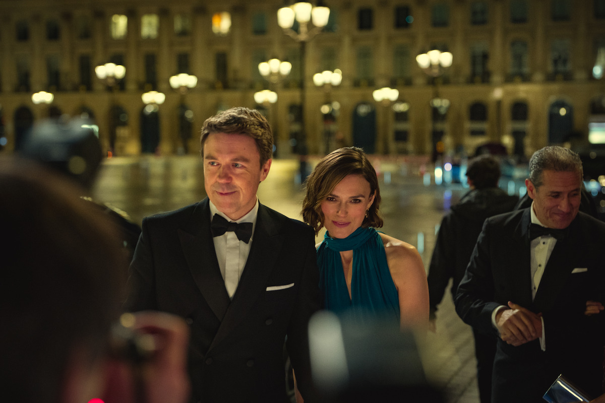 Um homem de smoking e uma mulher de vestido azul posam para fotos durante um grande evento noturno a céu aberto, com muitas luzes e um prédio majestoso ao fundo. Pessoas com trajes formais também aparecem por perto.