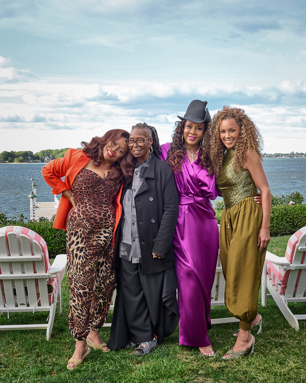 Four women pose together outdoors by a lake, standing on grass in front of white chairs. They are dressed in vibrant outfits, smiling, with a scenic water and cloudy sky backdrop.