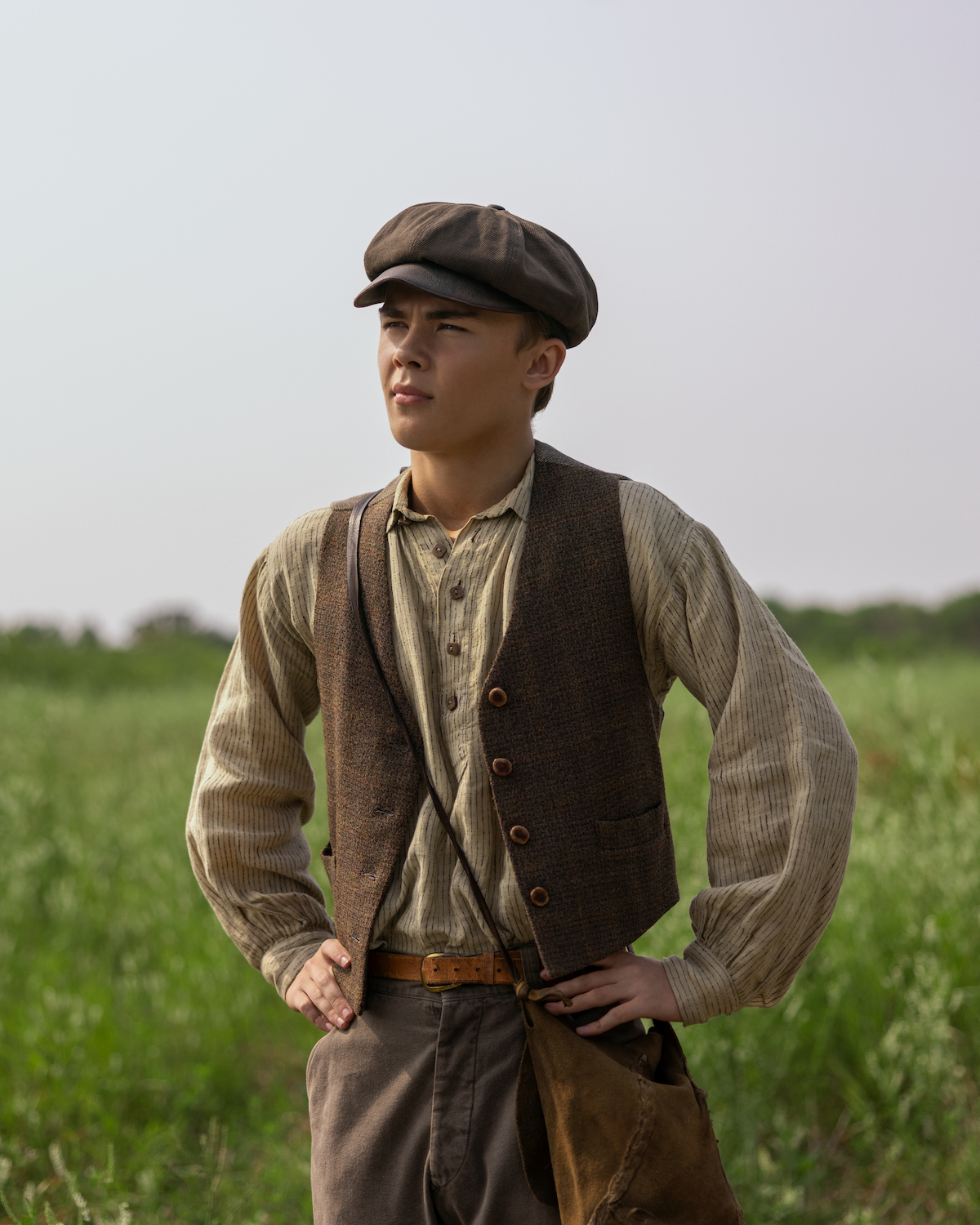 Young man wearing vintage clothes and a flat cap stands in a green field under a clear sky, hands on hips, carrying a brown bag, evoking an early 20th-century rural setting.