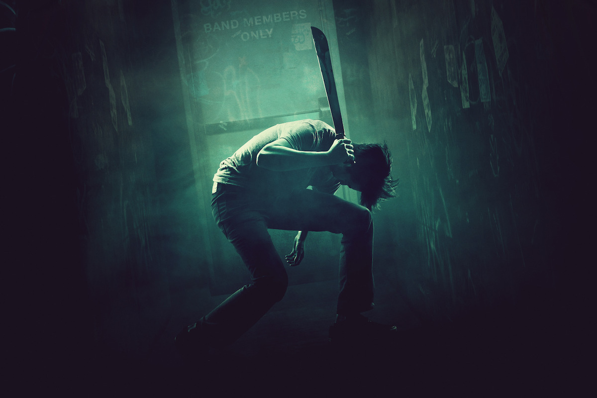 A person crouching in a dark, smoky hallway holding a machete, dramatic lighting casting shadows, posters on the walls, and a lit doorway labeled "Band Members Only" in the background.