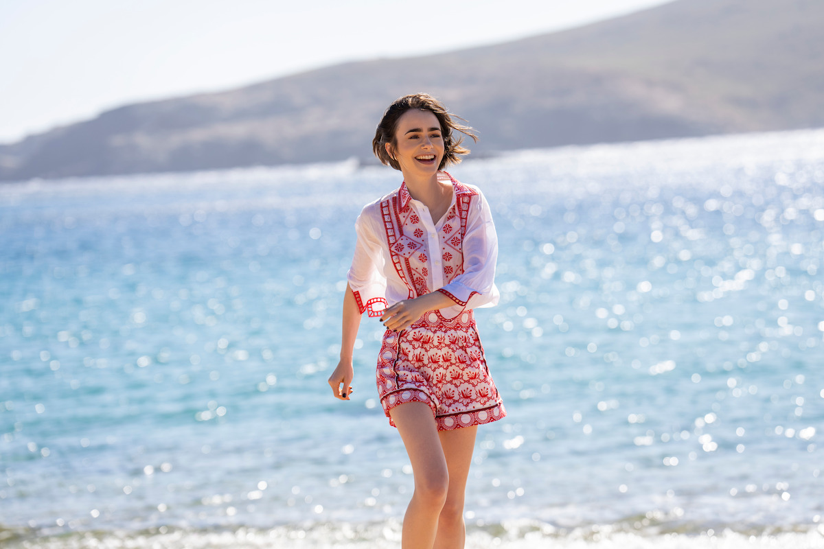 A young woman in a patterned shirt and shorts smiles while walking along a sunny beach, with sparkling blue water and distant hills in the background.