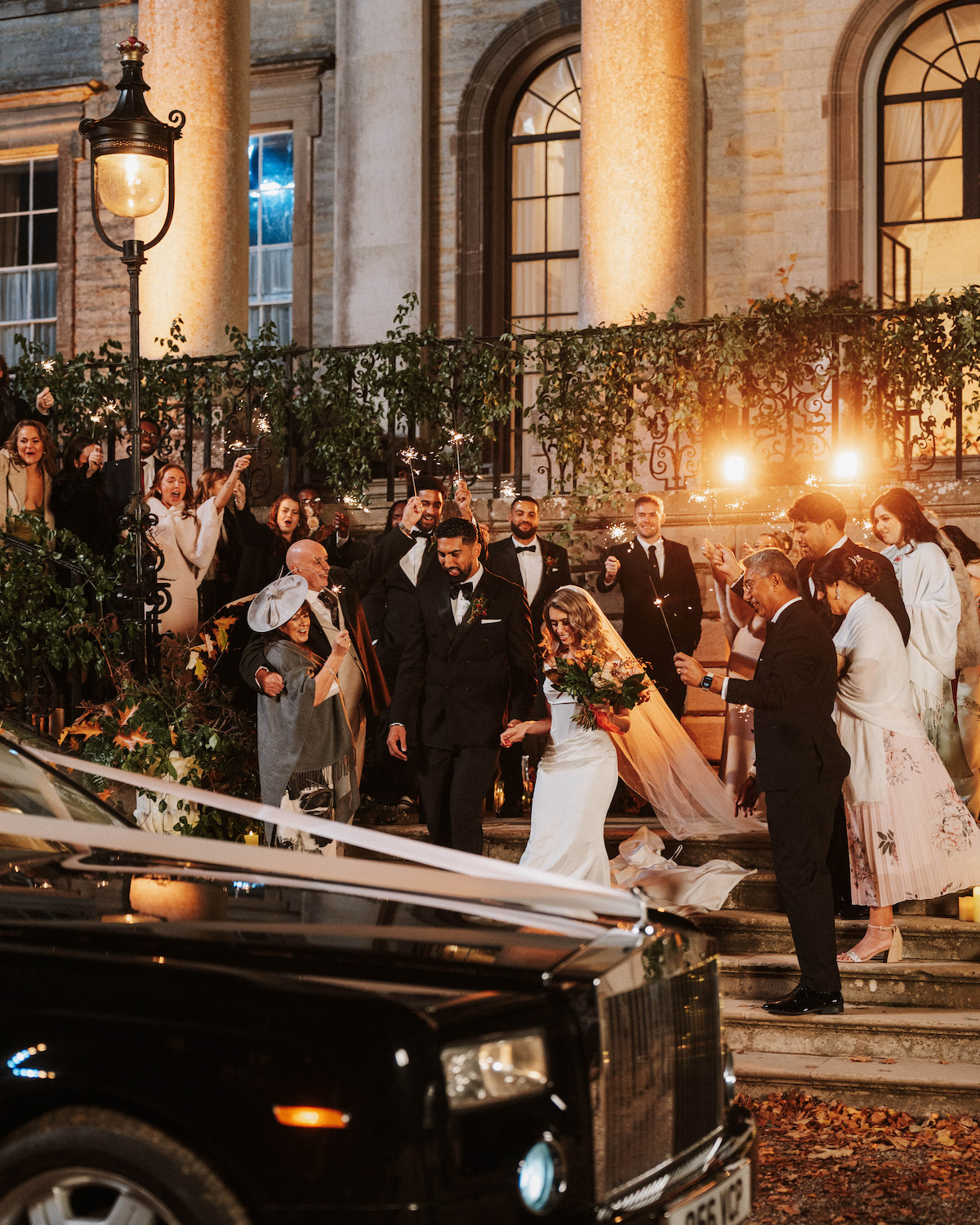 Bride and groom exit grand building at night, surrounded by cheering guests on steps, greenery, warm lights, and a black car with white ribbons in the foreground, setting a festive wedding celebration atmosphere.