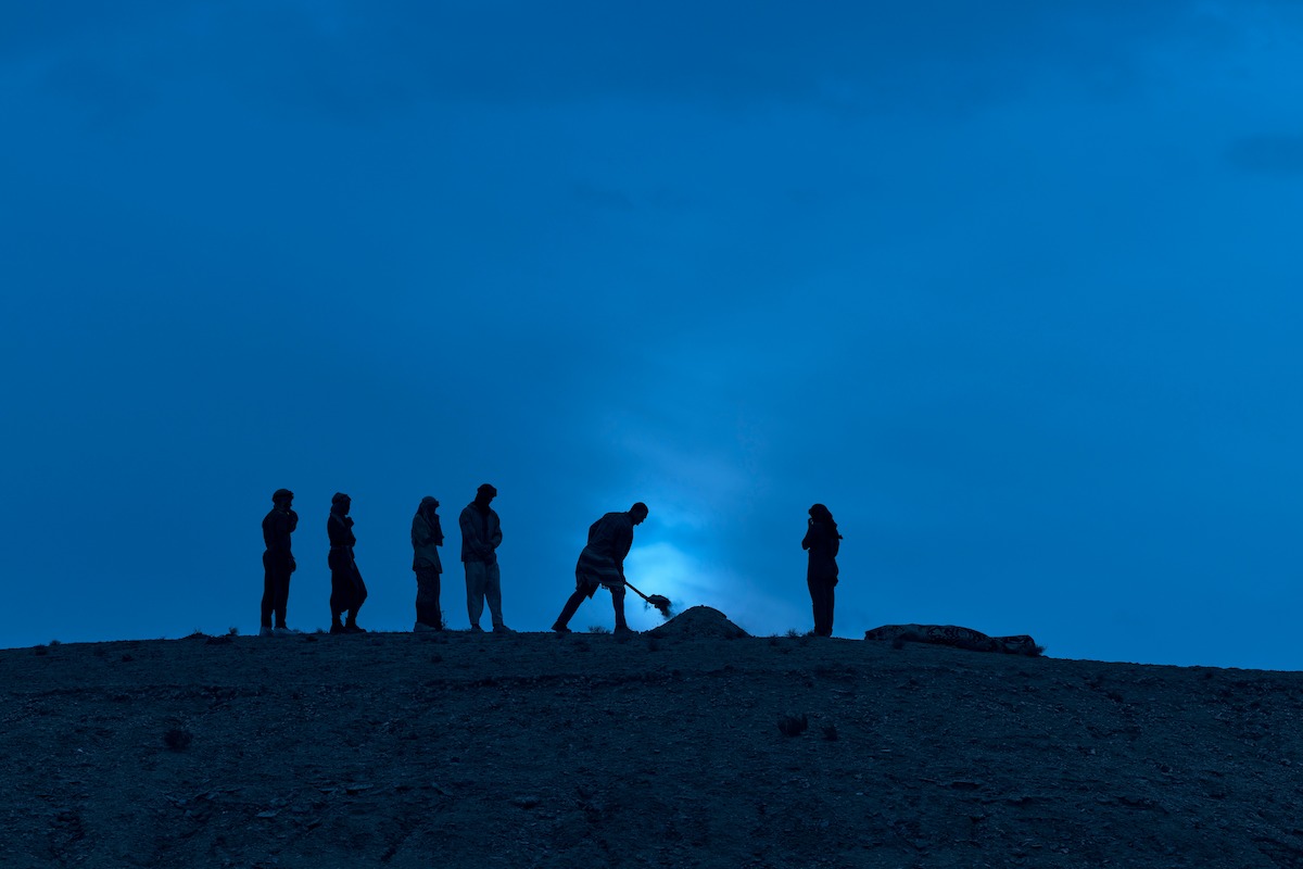 Silhouettes of six people standing on a hill at dusk, with one person digging, under a blue sky.