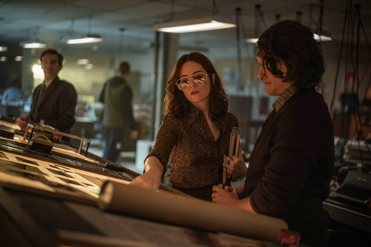 A woman in 1970s dress and large glasses talks with a colleague over a layout table in a dim newsroom.