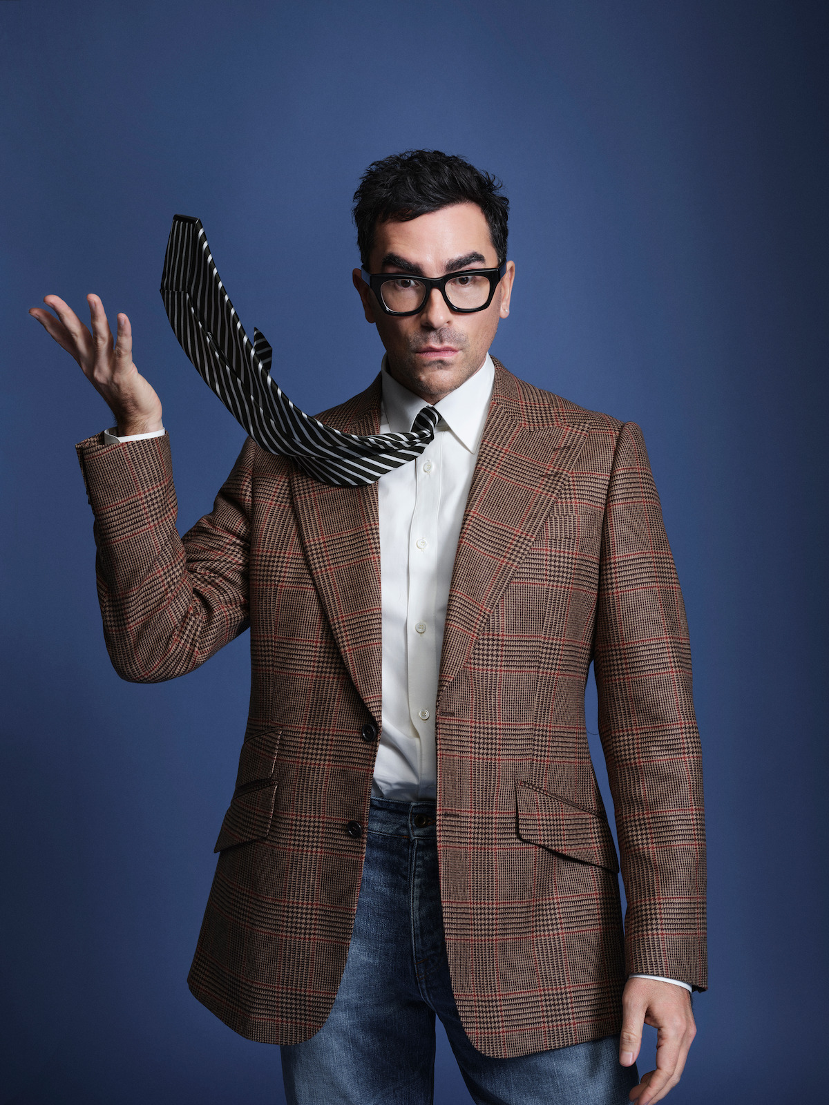 Dan Levy poses against a blue backdrop, wearing a checked blazer.
