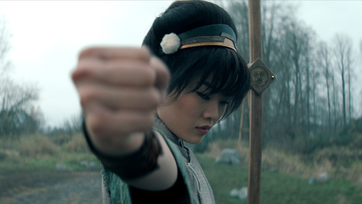 Miyako as Toph in traditional attire strikes a martial arts pose outdoors, focusing forward with a determined expression, holding a staff, with a backdrop of trees and cloudy sky in a natural, open setting.
