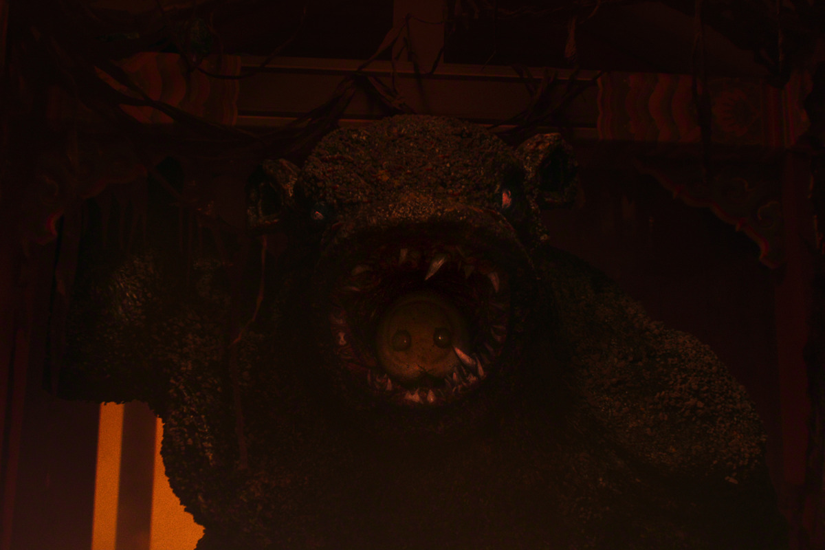 A monstrous, bear-like creature with sharp teeth stands in a dark, eerie environment, illuminated by dim, warm light with tattered decorations hanging above, creating a menacing and sinister atmosphere.