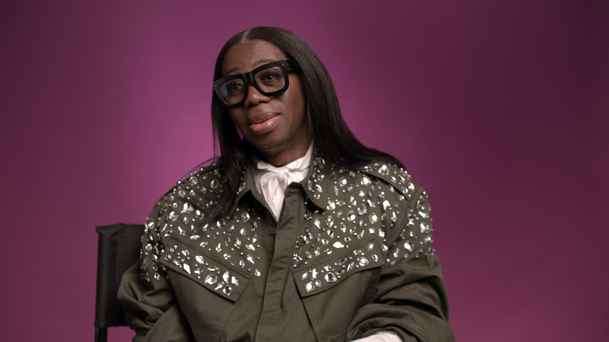 Person with long hair and glasses, wearing a studded olive jacket and white shirt, sits on a chair against a plain purple background in a studio setting.
