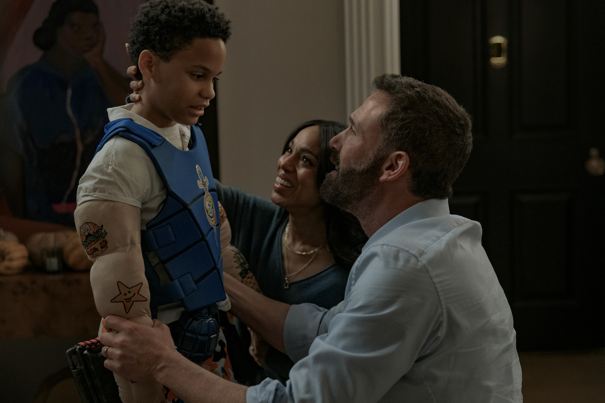 A child wearing a muscle costume and blue vest stands while smiling adults, a man kneeling and a woman crouching, look at him warmly in a cozy, dimly lit room with artwork and furniture in the background.