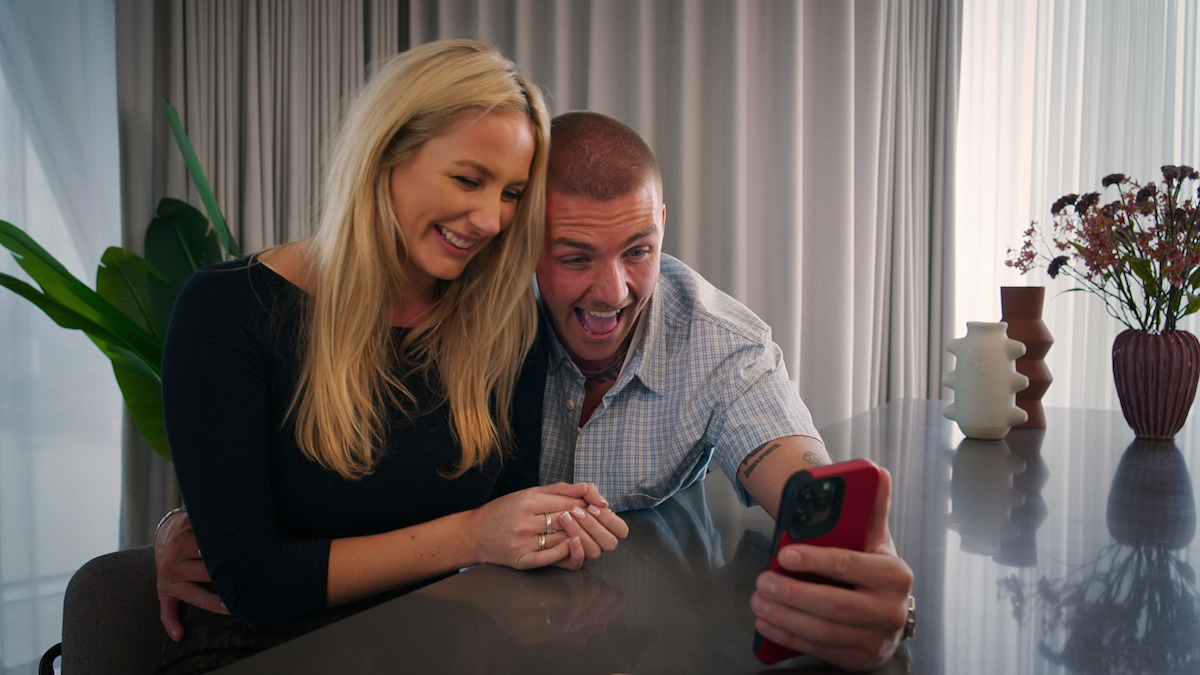 Smiling couple sitting at a table indoors, taking a selfie or video call on a smartphone, with modern decor, plants, and vases in the background.
