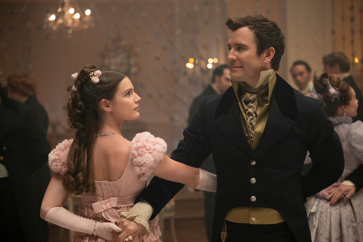 A man and woman in elegant period costumes dance at a formal ball, surrounded by other couples in a grand, chandelier-lit ballroom with soft lighting and festive decorations.