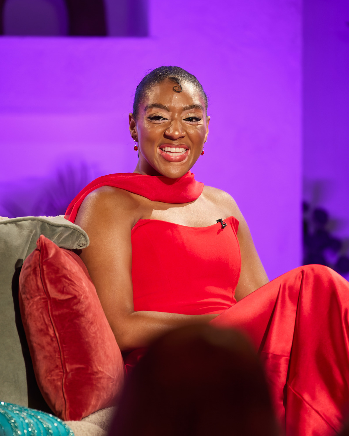 Woman in a bright red dress sitting on a couch with red and blue pillows, smiling in a warmly lit indoor setting with purple background lighting, suggesting a relaxed or interview environment.
