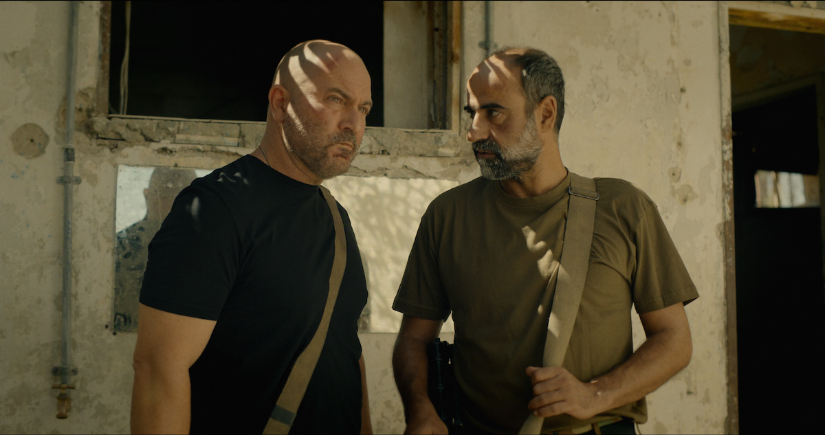 Two serious men with shoulder bags stand in front of a weathered, sunlit building, partially shaded by window light inside a worn, possibly abandoned environment.