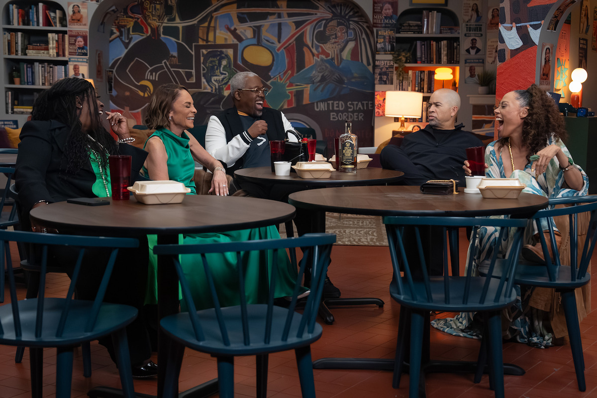 Five people sit around tables in a colorful cafe or library setting, laughing and conversing with takeaway food containers and drinks, surrounded by bookshelves and mural art in the background.