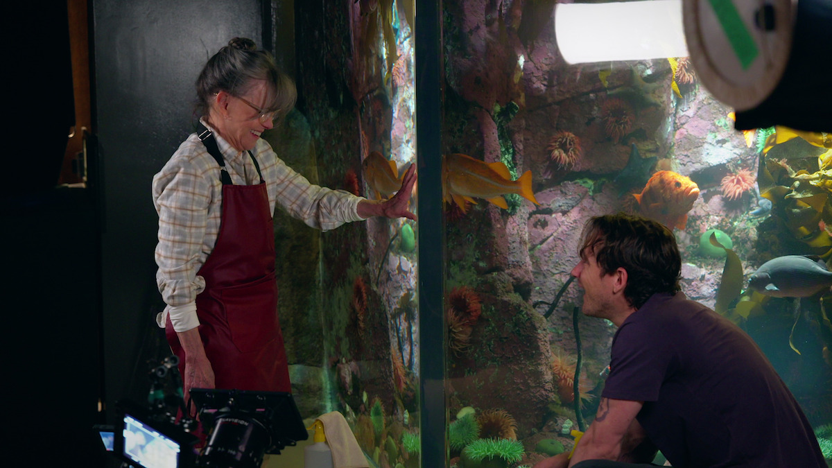 Sally Field as Tova and Lewis Pullman as Cameron engage at a large aquarium with colorful fish and sea life, surrounded by studio lights and filming equipment, suggesting a behind-the-scenes setting at an aquarium exhibit.
