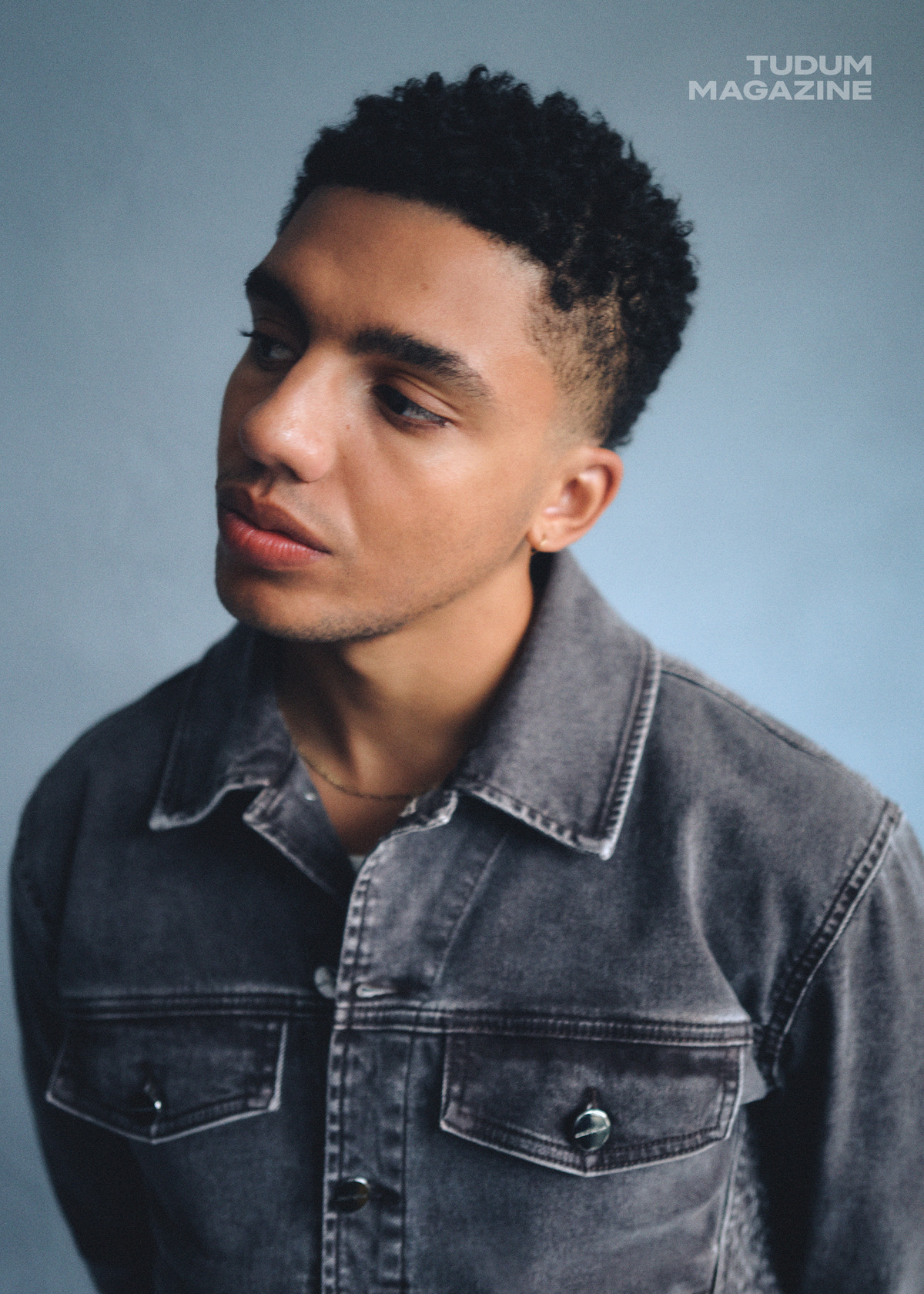 Jay Lycurgo stands against a blue wall, looking off to the left pensively.