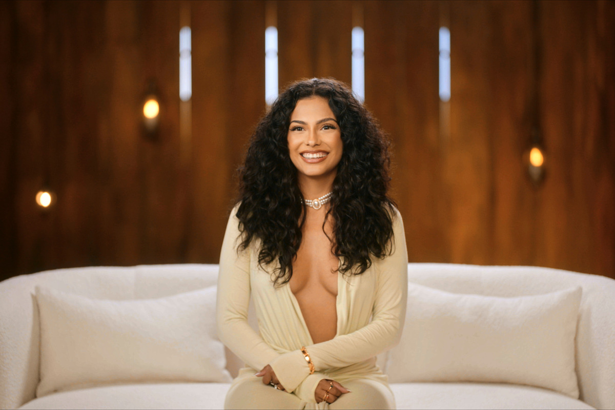 Woman with long curly hair in a cream outfit sitting on a white couch, smiling, in a warmly lit room with wooden walls and decorative lights in the background.