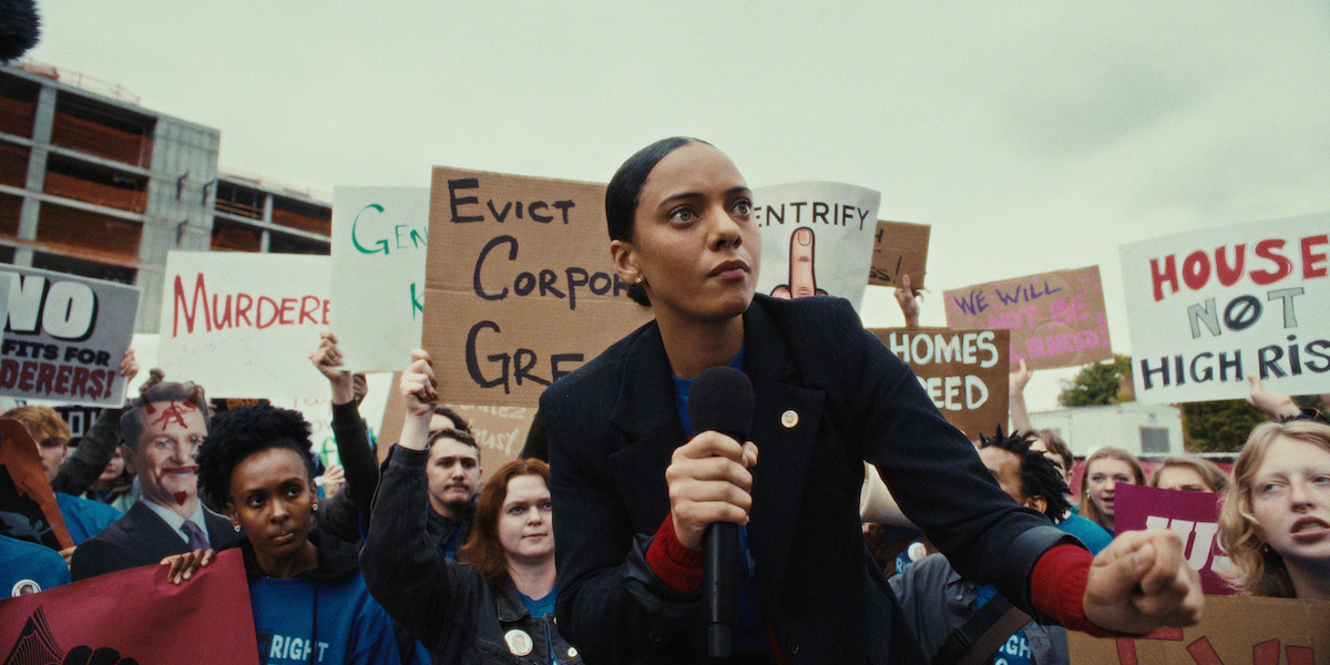 A serious woman with a microphone leads a protest outdoors, surrounded by diverse people holding signs against corporate greed and gentrification; the mood is intense and urgent, calling for social justice and housing rights.