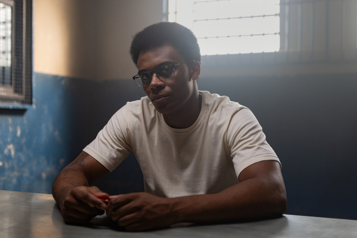 Um homem negro usando óculos e camisa branca se senta numa mesa dentro de um presídio