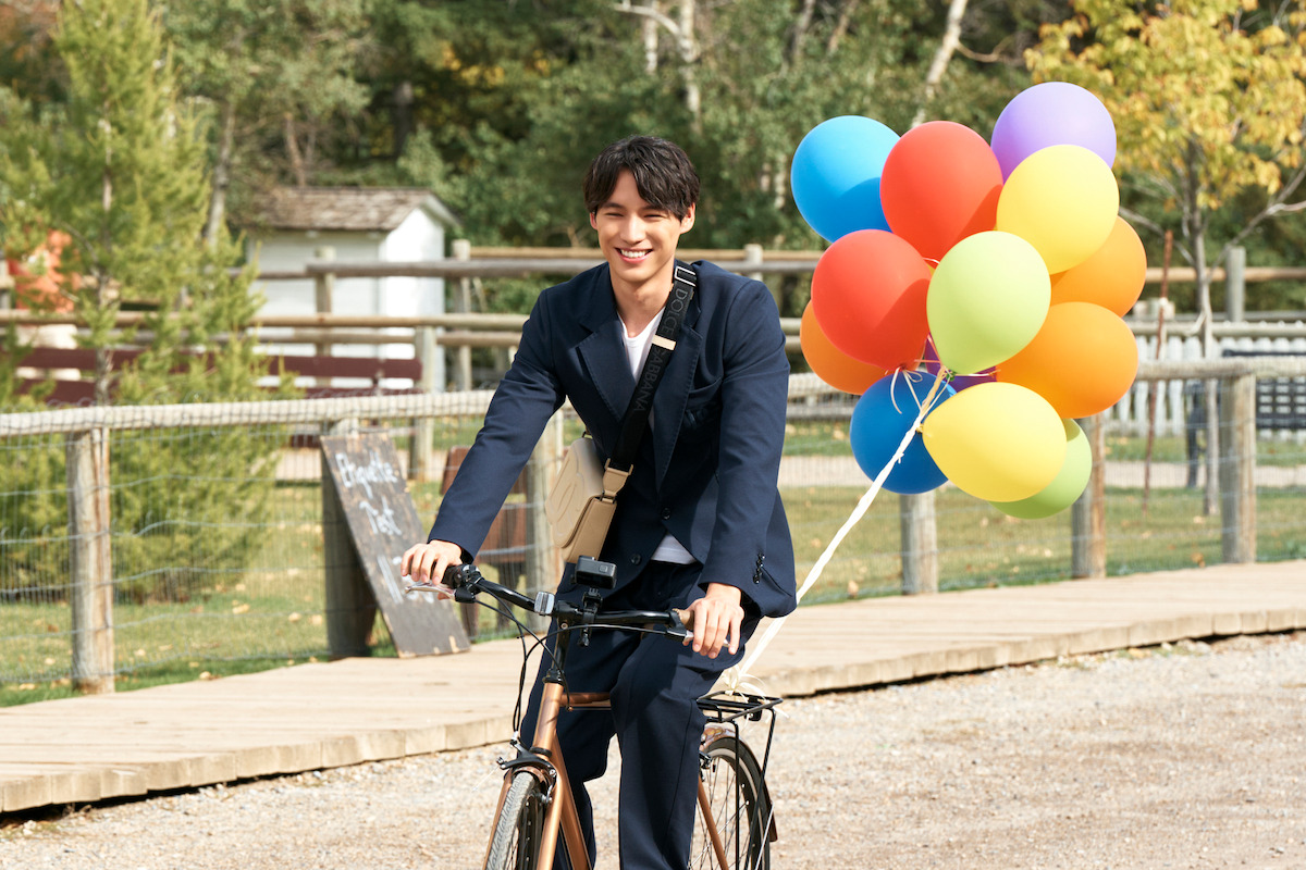 A young man in a suit rides a bicycle outdoors on a gravel path, holding colorful balloons, with green trees and a wooden fence in the background on a sunny day.