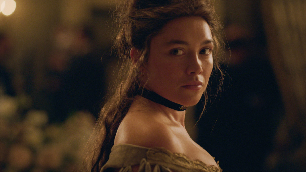 A woman in period costume with a black choker looks over her shoulder in a dimly lit, elegant room, possibly at a formal event or ball. The background is softly blurred with warm lighting and floral decorations.