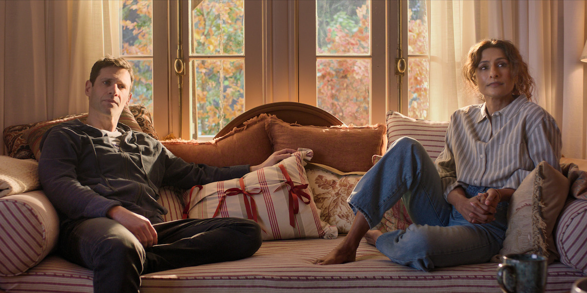A man and woman sit apart on a light-colored couch in a cozy living room with large windows, autumn trees outside, and soft sunlight filtering in. The mood is calm and contemplative.