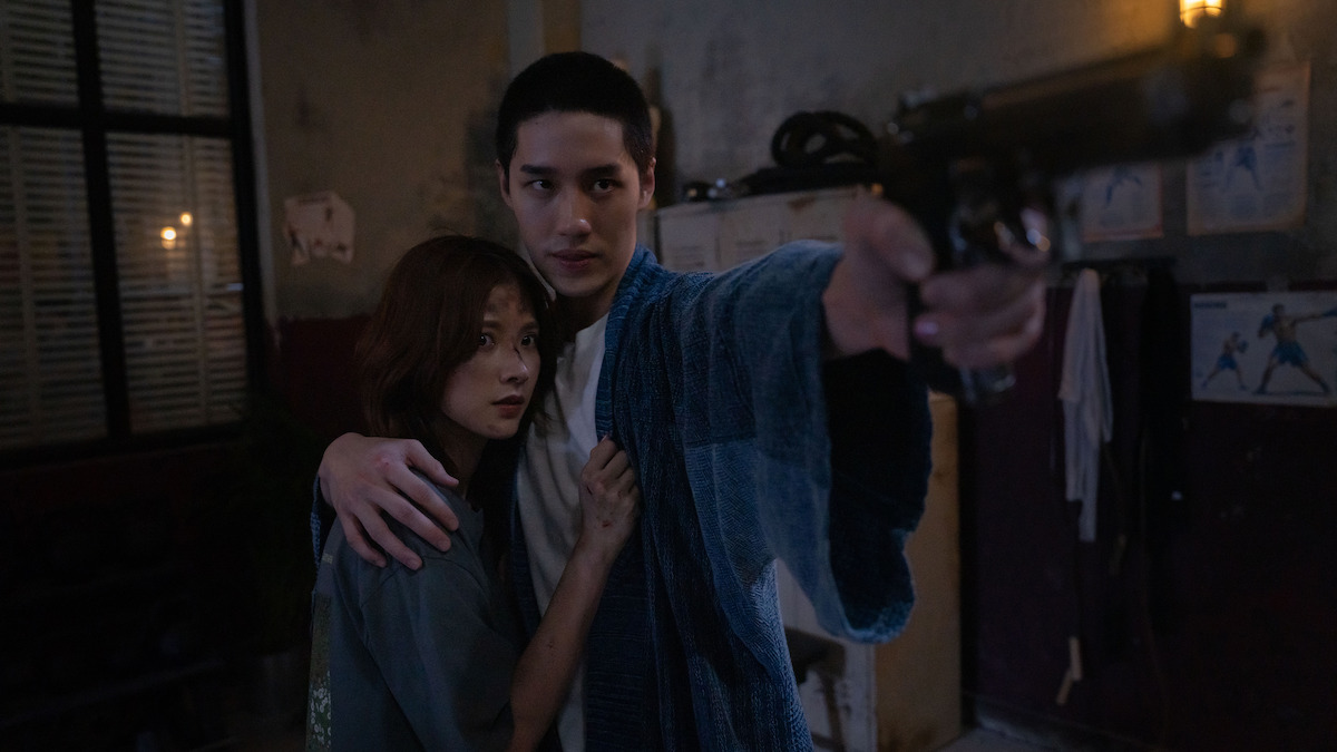 A man in a blue robe holds a woman protectively while aiming a handgun in a dimly lit, cluttered room with posters on the wall and barred windows.