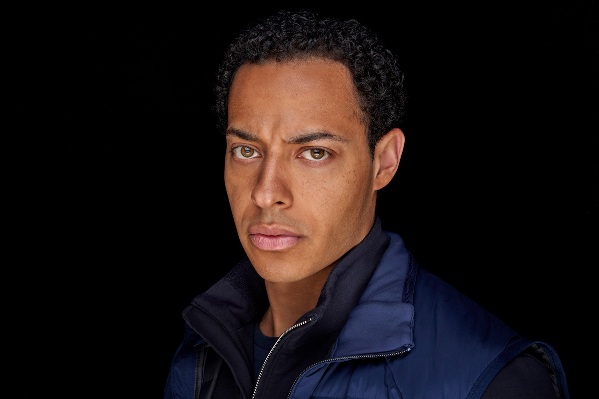 Man with short curly hair wearing a blue jacket, photographed against a solid black background. The lighting is dramatic, highlighting his serious facial expression and creating a focused, intense mood.