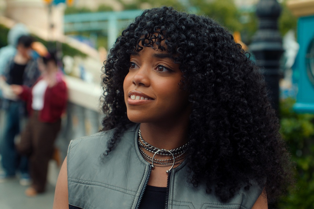 Young woman with curly hair smiling outdoors in an urban setting, wearing a gray vest and layered chain necklaces, with people and greenery blurred in the background.