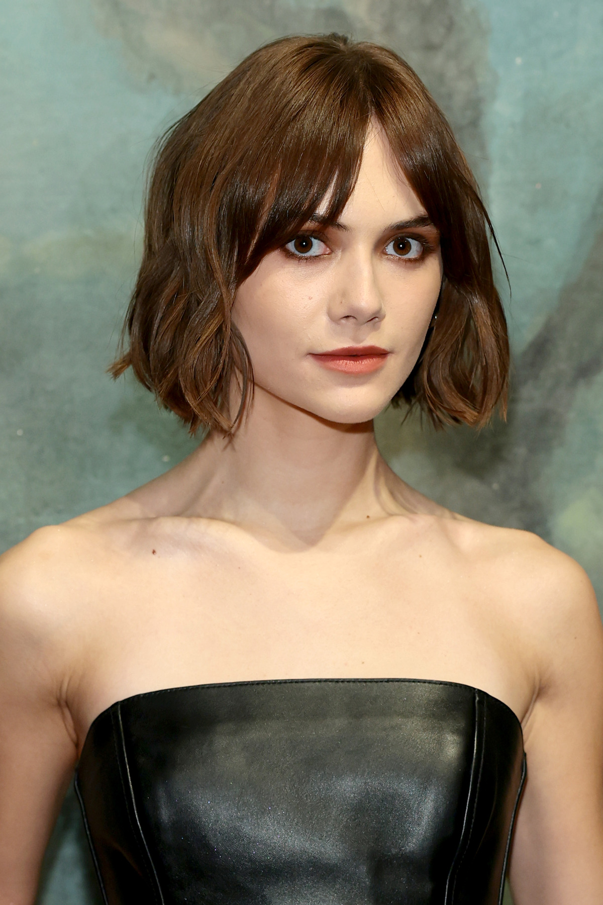 Woman with short brown hair wearing a strapless black leather dress, posing against a soft, painted backdrop in a formal setting, looking directly at the camera with a neutral expression.