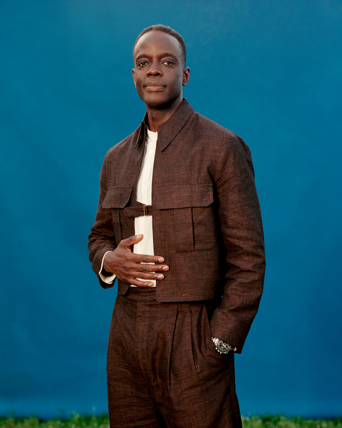 Ato Essandoh wearing a brown jacket and pants standing in front of a blue background.