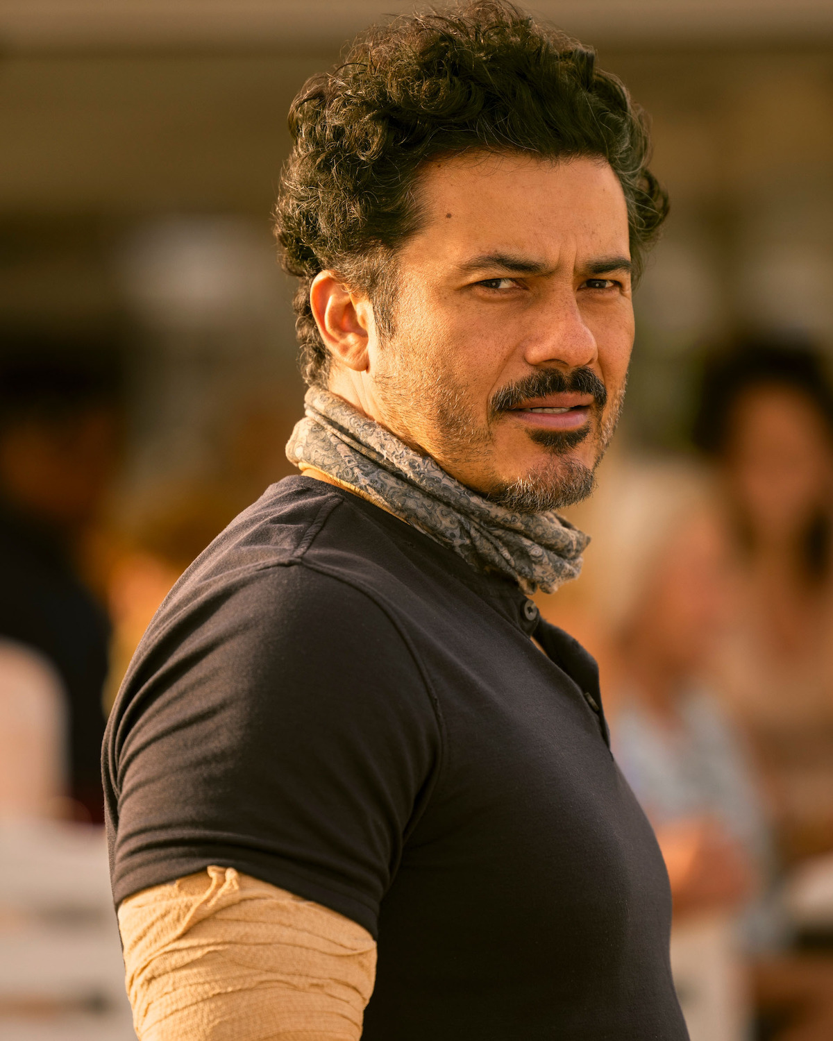 Rigo Sanchez as Lightner, with curly hair, beard, and mustache wearing a dark shirt and scarf, standing outdoors in warm lighting with blurred people and objects in the background, looking over his shoulder.